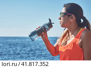 Latin woman, middle-aged, resting, regaining strength, eating, drinking water, after a gym session, burning calories, keeping fit, outdoors by the sea, wearing headphones and smart watch. Стоковое фото, фотограф carlos l vives / Фотобанк Лори