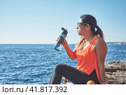 Latin woman, middle-aged, resting, regaining strength, eating, drinking water, after a gym session, burning calories, keeping fit, outdoors by the sea, wearing headphones and smart watch. Стоковое фото, фотограф carlos l vives / Фотобанк Лори