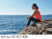 Latin woman, middle-aged, resting, regaining strength, eating, drinking water, after a gym session, burning calories, keeping fit, outdoors by the sea, wearing headphones and smart watch. Стоковое фото, фотограф carlos l vives / Фотобанк Лори