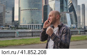 Счастливый молодой человек, разговаривает по телефону, радуется успехам или хорошим новостям. Красивый мужчина прогуливается по набережной Москвы-реки на фоне бизнес-центра Москва-сити летним, теплым днем. Симпатичный лысый парень общается  по смартфону. Стоковое видео, видеограф Антон Слабоспицкий / Фотобанк Лори