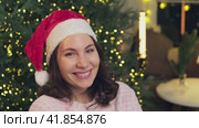 Portrait of a young attractive woman in a red Christmas hat sitting in an armchair in cozy pajamas, embarrassed, looking at the camera, turning her head and smiling. Christmas and New Year holidays. Стоковое видео, видеограф Денис Кебедов / Фотобанк Лори