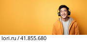 Portrait of happy laughing asian young man in headphones on yellow orange. Banner. Copy space. Стоковое AI-изображение, AI-художник Наталья Семкина / Фотобанк Лори