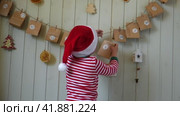 Boy in pajamas, Santa cap eating sweet candy on paper advent calendar with presents background. Little happy child with gift of handmade Christmas calendar on wall. Celebrating at home Xmas tradition. Стоковое видео, видеограф Марина Демидюк / Фотобанк Лори