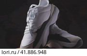 Two white women's sneakers on a wooden surface. Dark background. The camera flies around. Parallax effect. Close-up. Krasnodar, Russia, April 19, 2022. Редакционное видео, видеограф Воловодов Владимир / Фотобанк Лори