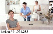 Sad offended young woman sitting at kitchen table during argument with dissatisfied husband and mother-in-law standing behind on blurred background. Concept of family fights. Стоковое видео, видеограф Яков Филимонов / Фотобанк Лори