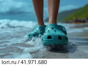 Crocs shod feet of an person on the beach walking along the surf. Стоковое AI-изображение, AI-художник Евгений Харитонов / Фотобанк Лори
