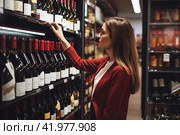 A young woman is shopping at the supermarket, buying groceries, choosing wine, Alcohol, holding a bottle. People buying the gastronomic concept of food. Стоковое AI-изображение, AI-художник Ирина Ручушкина / Фотобанк Лори