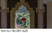 An elegant arched window opening in the Kek Lok Si Temple. A bright red Chinese lantern is suspended against a background of lush green vegetation. Malaysia. Penang. (2023 год). Стоковое фото, фотограф Вера Ивановна Кубарева / Фотобанк Лори