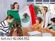 Female teacher, conducting extracurricular lesson in the school class tells schoolchildren the history of Mexico and shows national flag of country. Стоковое фото, фотограф Яков Филимонов / Фотобанк Лори