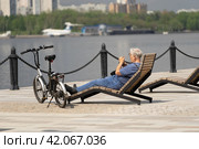 Moscow, Russia - May, 15, 2021: The eldest man alone in the summer sits on the embankment of the river and looks at the phone. He's resting after a bike ride. Concept of active sports life in old age. Редакционное фото, фотограф Валерий Леонидович / Фотобанк Лори