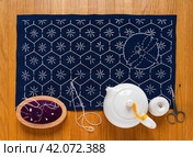 Flat lay of process of hand embroidery in Sashiko style on blue canvas with white threads of geometric patterns and dragonfly. Embroidery supplies and teapot with green tea on wooden table. Close-up, top view. Стоковое фото, фотограф Виктория Катьянова / Фотобанк Лори