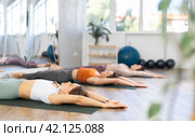 Sporty women doing yoga with group in studio, lying on mat with knee pressed to chest with hands, performing hamstring and low back stretching Ardha Pawanmuktasana. Стоковое фото, фотограф Яков Филимонов / Фотобанк Лори
