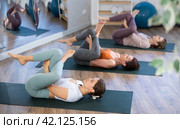 Sporty women doing yoga with group in studio, lying on mat with knee pressed to chest with hands, performing hamstring and low back stretching Ardha Pawanmuktasana. Стоковое фото, фотограф Яков Филимонов / Фотобанк Лори