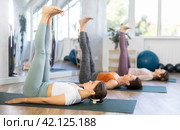Sporty women doing yoga with group in studio, lying on mat with knee pressed to chest with hands, performing hamstring and low back stretching Ardha Pawanmuktasana. Стоковое фото, фотограф Яков Филимонов / Фотобанк Лори