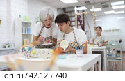 Experienced mature female ceramicist teaching young enthusiastic guy art of ceramic glazing and painting in modern pottery studio. Стоковое видео, видеограф Яков Филимонов / Фотобанк Лори
