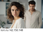 Upset woman with curly hair standing in a kitchen looking at the camera with a man in the background. Family conflict. Стоковое AI-изображение, AI-художник Оксана Гильман / Фотобанк Лори