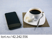 Белая чашка чёрного кофе с сахаром на салфетке-циновке и телефон на столе ранним утром. Стоковое фото, фотограф Владимир Устенко / Фотобанк Лори