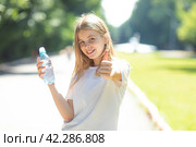 Portrait of beautiful teenager pretty girl, young thirsty woman is drinking pure fresh water from bottle at warm sunny summer day outdoors, holding bottle in hand and showing thumbs up. Стоковое фото, фотограф Ирина Михайличенко / Фотобанк Лори
