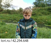Sremska Mitrovica, October 18, 2021. A blond boy of 8 years old walks near a swampy pond in autumn. Warm green clothes. The boy grimaces. Emotion of resentment. The man bulges the sluice. Редакционное фото, фотограф Юлия Петровская / Фотобанк Лори