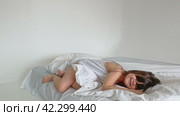 A nice child girl enjoys sunny morning. Good morning at home. Child girl wakes up from sleep. Sweet little girl is looking at camera and smiling while sitting on her bed on white background. Стоковое видео, видеограф Ирина Михайличенко / Фотобанк Лори