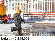 Portrait of a little boy in a yellow hat. The child runs through puddles on the farm, has fun and plays. It's a sunny day. Early spring. The first drop. Стоковое фото, фотограф Photojuli / Фотобанк Лори