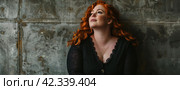 Beautiful size-plus redhead curly-haired woman posing on grunge old-fashioned wall background. Стоковое AI-изображение, AI-художник Alexander Tihonovs / Фотобанк Лори