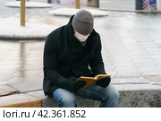 Handsome man in Moscow city center during coronavirus pandemic. New Year holidays. He sitting at the bench and reading the book in the public park. Стоковое фото, фотограф YuryKara / Фотобанк Лори