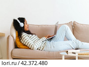 African-American female in headphones surfing the net on laptop while lying on sofa in living room. Стоковое фото, фотограф Natalia Gdovskaia / Фотобанк Лори