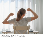 Young female stretching while sitting at desk at home. Remote wo. Стоковое фото, фотограф Natalia Gdovskaia / Фотобанк Лори