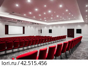 Conference room with red chairs, interior lighting, and empty white insulated screen. Business meetings, conferences, trainings, used as template for elegant design. Interior of the conference hall. Стоковое AI-изображение, AI-художник Aleksandr Golubev / Фотобанк Лори