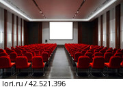 Conference room with red chairs, interior lighting, and empty white insulated screen. Business meetings, conferences, trainings, used as template for elegant design. Interior of the conference hall. Стоковое AI-изображение, AI-художник Aleksandr Golubev / Фотобанк Лори