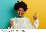 ctp afroamerican young woman in white sweater with thumbs up while pointing to right. Стоковое AI-изображение, AI-художник Юлия Гапеенко / Фотобанк Лори