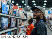 Employee wearing a cap and glasses in front of electronics store shelves. Стоковое AI-изображение, AI-художник Юрий Поздников / Фотобанк Лори