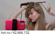 A woman styles hair using a curling wand, manual wrapping of long blonde hair around the wand to create curls. Blondie is live streaming the process on smartphone (2024 год). Редакционное видео, видеограф А. А. Пирагис / Фотобанк Лори