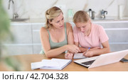 Positive woman offering assistance and support to teenage daughter absorbed in writing notes in study book while sitting at table with laptop. Стоковое видео, видеограф Яков Филимонов / Фотобанк Лори