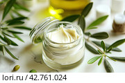 Cosmetic skin cream on a white table surrounded by fresh green leaves and olive oil. Стоковое AI-изображение, AI-художник Ярославский Максим / Фотобанк Лори