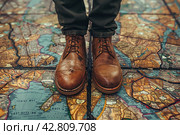 Legs of a man in stylish brown leather lace-up shoes standing on a surface covered with a large map. The map appears to be ancient, with images of lands, bodies of water and names in Latin, done in a traditional style. Стоковое AI-изображение, AI-художник Анна Кривицкая / Фотобанк Лори