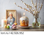 Orthodox Easter. Easter cake, painted eggs, icons, faces of saints, flowers, church candles and Easter decor on a wooden table in an old village house. Стоковое AI-изображение, AI-художник Photojuli / Фотобанк Лори