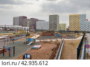 Благоустроенная детская игровая зона, огороженная металлической изгородью-забором на фоне многоэтажных разноцветных жилых домов новостроек. Горка, лестница, песочница, беседка. Пасмурное небо с облаками, слегка пробиваются лучи солнца. (2024 год). Редакционное фото, фотограф Сергей Никифоров-Гордеев / Фотобанк Лори