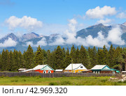 Village wooden houses against backdrop of spruce forest and Eastern Sayan Mountains with low clouds on Tunka foothill valley on sunny summer day. Scenic rustic landscape. Siberia. Buryatia. Arshan. Стоковое фото, фотограф Виктория Катьянова / Фотобанк Лори