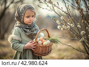 Happy bright Easter. Christianity. Portrait of a three-year-old girl in a Russian folk dress and shawl with a wicker basket with Easter eggs in her hands against a background of a flowering willow. Стоковое AI-изображение, AI-художник Photojuli / Фотобанк Лори