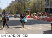 В Москве по улице Академика Комарова мимо бригады рабочих, меняющих бордюрный камень на тротуарах, едет на электровелосипеде курьер в обычной одежде с голубыми пакетами, торчащими из корзины и свисающими с руля (2024 год). Редакционное фото, фотограф Наталья Николаева / Фотобанк Лори
