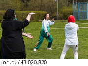 Молодая женщина инструктор в китайском спортивном костюме демонстрирует перед учениками выполнение движений руками. Занятие цигун на зеленой поляне в городском парке. (2024 год). Редакционное фото, фотограф Сергей Никифоров-Гордеев / Фотобанк Лори
