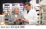 Concerned old female customer asking middle-aged chemist about product reflected on her phone in large pharmacy. Стоковое видео, видеограф Яков Филимонов / Фотобанк Лори