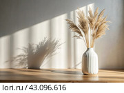 A modern white ceramic vase with dried grass on a wooden table. Scandinavian interior. An empty white wall with a shadow from the window. Стоковое AI-изображение, AI-художник Photojuli / Фотобанк Лори