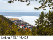 Посёлок Листвянка, Иркутская область, Россия. Вид сверху на дома и колесо обозрения на берегу озера Байкал. На переднем плане ветви деревьев. Вдали покрытое льдом озеро и горы. Весенний сезон. Май. (2023 год). Стоковое фото, фотограф Андрей Степанов / Фотобанк Лори