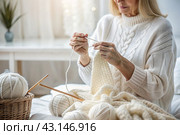 Close-up. A woman with balls of yarn knits a warm sweater in white and beige tones in her hands. Needlework, knitting. Winter season, small business, handmade clothes. Стоковое AI-изображение, AI-художник Photojuli / Фотобанк Лори