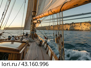 A boat under sail sails along the rocky seashore at dawn. Стоковое AI-изображение, AI-художник Ярославский Максим / Фотобанк Лори