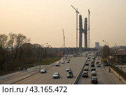 Вид сверху на автомобильную дорогу. Вдали строительство Центрального моста в Новосибирске. Башенные краны около строящейся высокой опоры вантового моста через реку Обь. Автомобили едут по широкой улице. Большевистская улица, город Новосибирск, Новосибирская область, Сибирь, Россия. (2023 год). Редакционное фото, фотограф Андрей Степанов / Фотобанк Лори