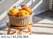 Balls of yarn in a wicker basket on a wooden stool. Needlework, textiles, knitting. Winter season, small business, handmade clothes. Стоковое AI-изображение, AI-художник Photojuli / Фотобанк Лори